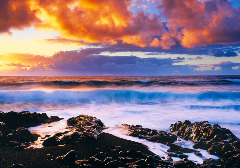 Waves crashing onto a rocky beach in Hana Hawaii with the sun rising into a cloud filled sky