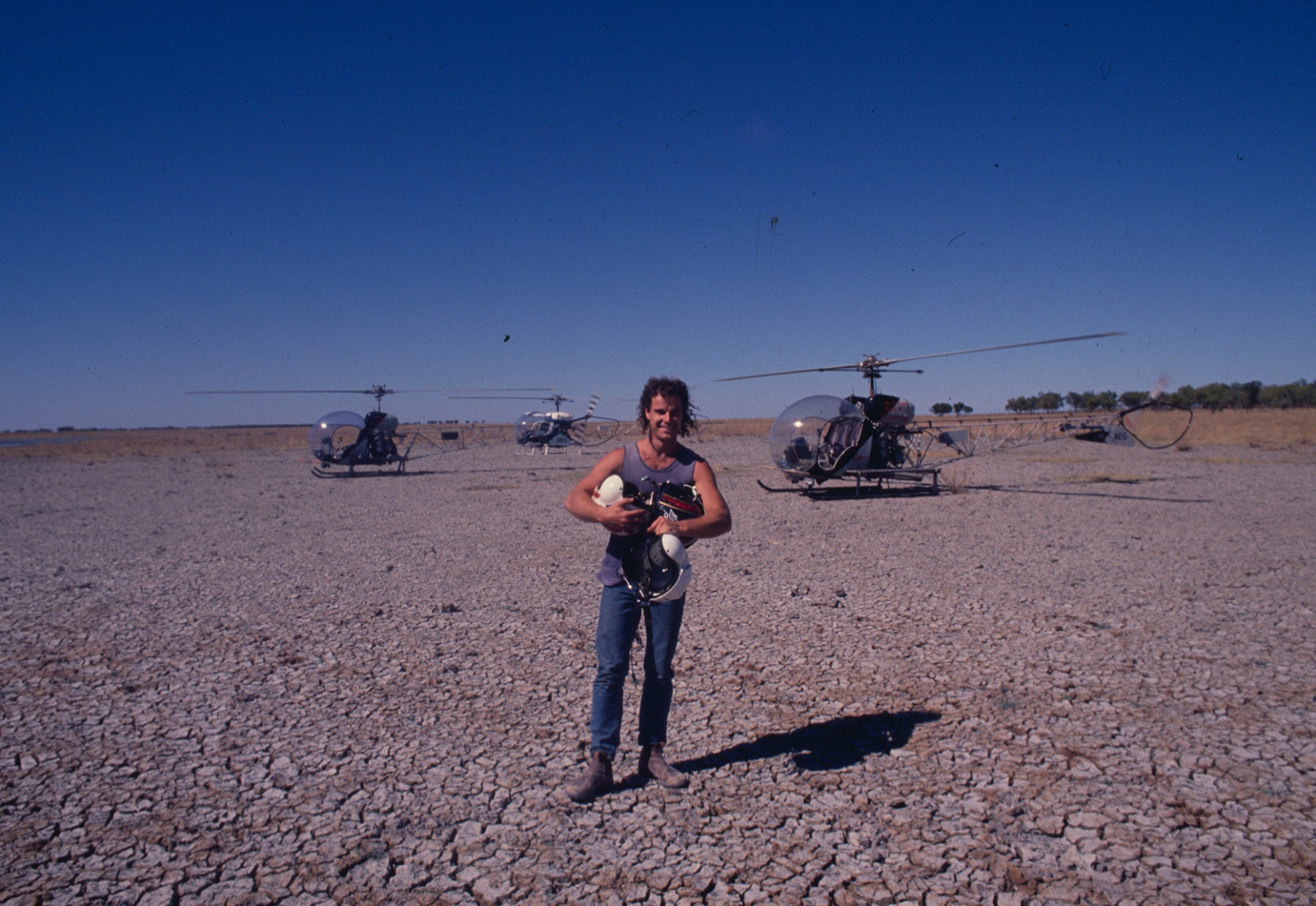 Early photograph of Peter Lik standing in a cracked, dry mud field in front of three helicopters holding a flight helmet
