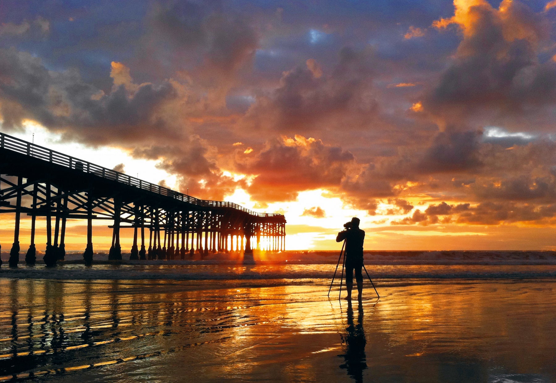 Silhouetted portrait of Peter Lik taking a photo of the Pacific Beach Pier in California at sunset.