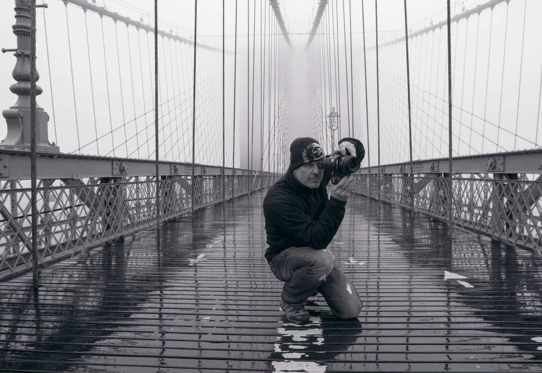 Black and white portrait of Peter Lik kneeling on the Manhattan Bridge during the rain taking a photograph with the steel support cables suspended in the background