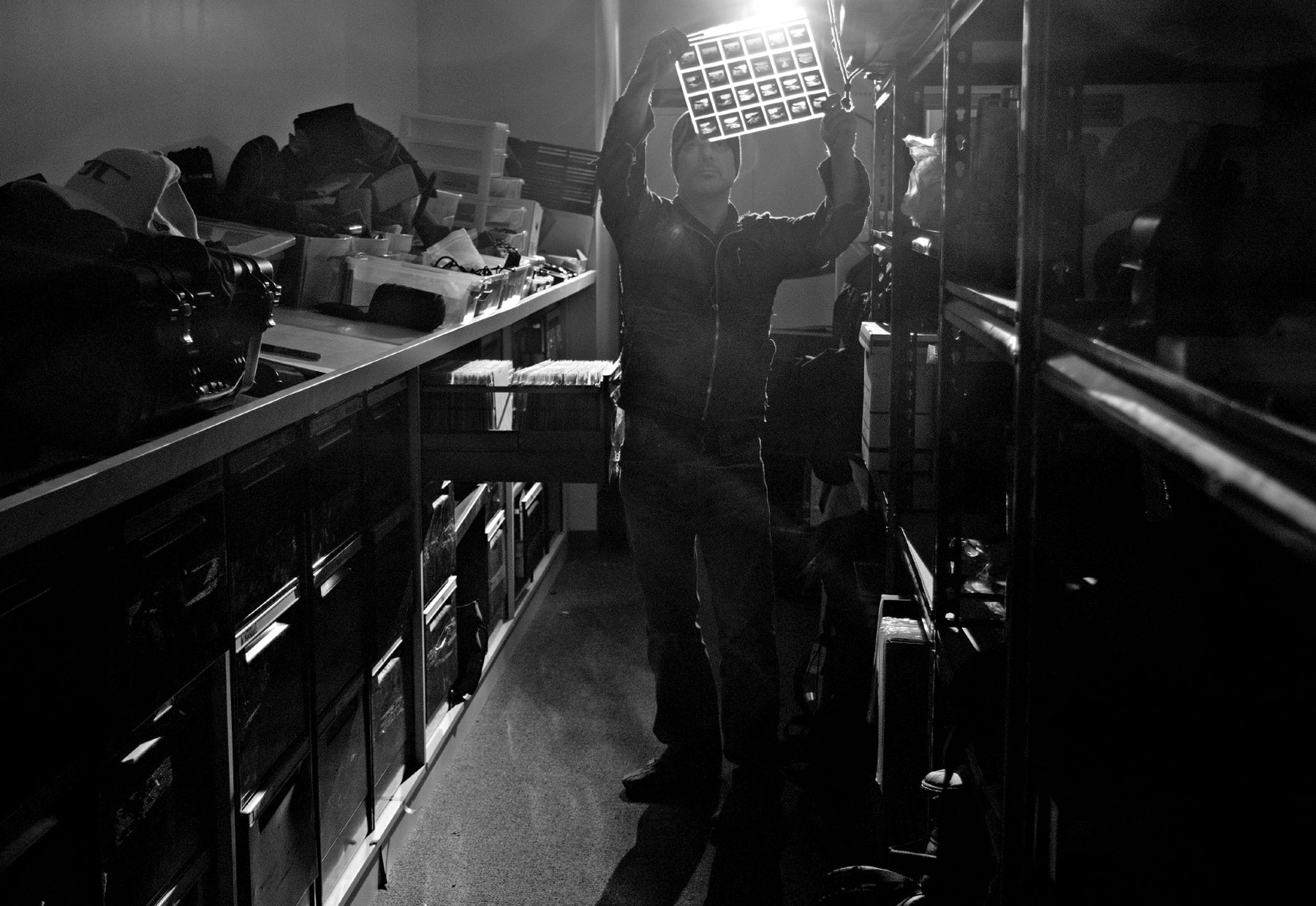 Backlit black and white portrait of Peter Lik wearing a black leather jacket and beanie viewing a sleeve of transparency slides standing in storage closet