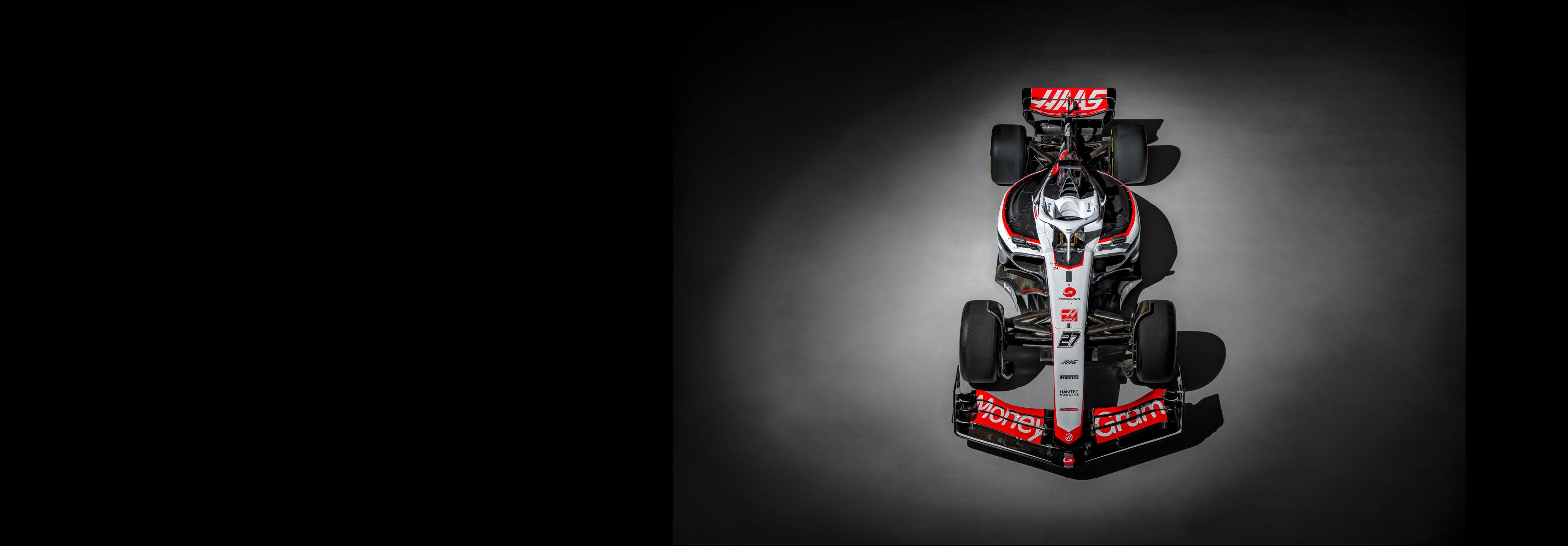 Photograph of the red, white, and black Haas F1 race car from above lit by a spotlight on the concrete floor of a airplane hanger.