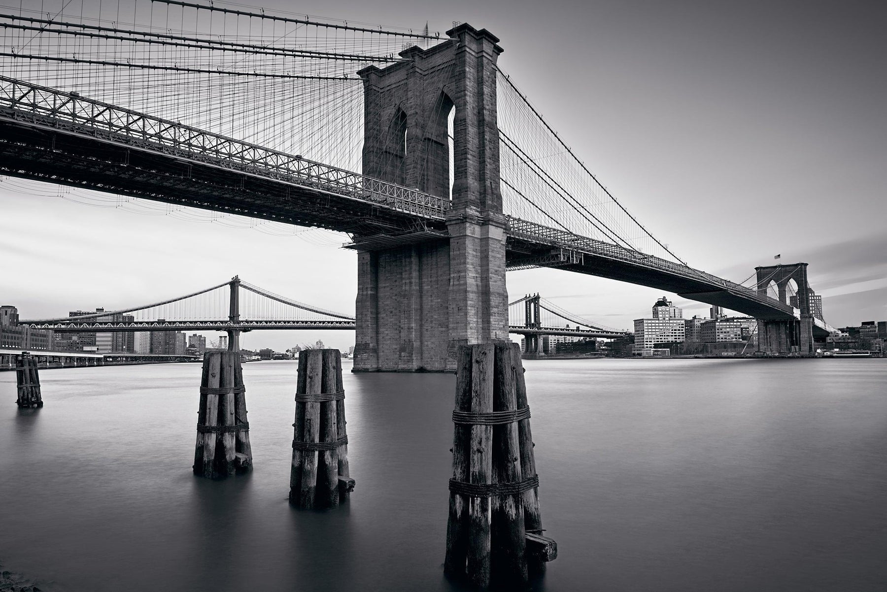 Black and white Brooklyn Bridge from the waters edge with the Manhattan Bridge and New York City in the background