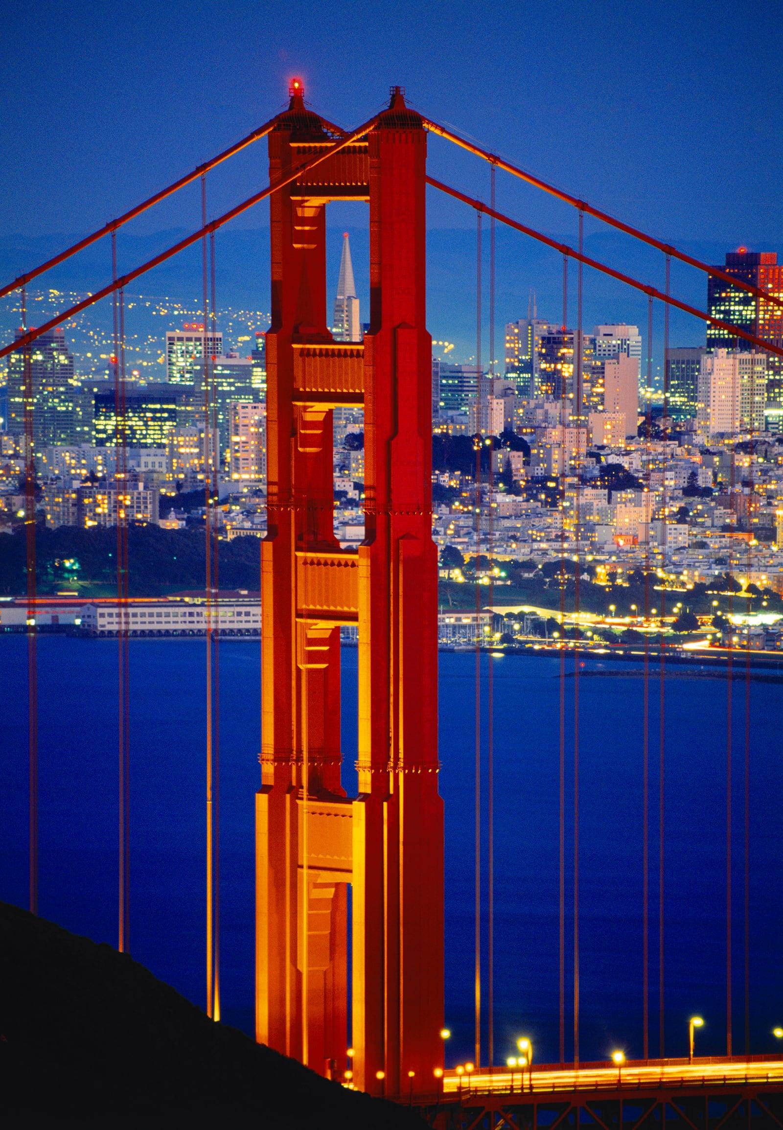 Close up of one of the Golden Gate Bridge towers at night with San Francisco lit up in the background