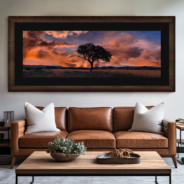 A brown wood framed photograph with a black liner hangs over a brown leather sofa with white pillows. The photograph is of a lone tree in a grass field in southern France. Clouds roll in on the blue skies as the sun sets below the low mountain range in the background.