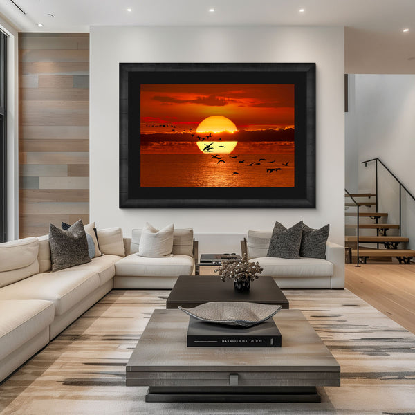A black framed photograph with a black liner hanging over a white sofa in a living room. The photo is of the bright yellow sun setting on the horizon of the ocean off the coast of Africa. Birds take fly and cast their silhouette in front of the large yellow sun. 