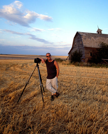 Portrait of Peter Lik standing in front of barn with his tripod in a grassy field.