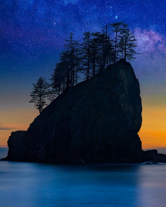 Silhouette of a sea stack covered with trees reflecting off the beach at sunset with the stars and milky way behind