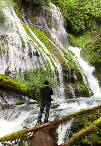 Portrait of Peter standing on a log in front of the towering waterfalls and moss covered rocks in  Gifford Pinchot State Park in Oregon