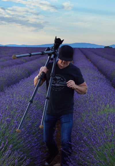 Portrait of Peter Lik wearing a black baseball cap and black t-shirt walking out of the purple Lavender of Valensole, France with a tripod over his shoulder