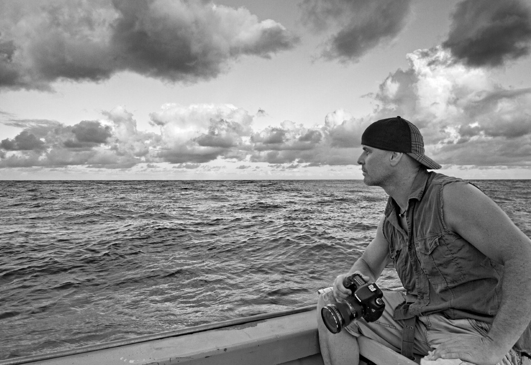 Black and white portrait of Peter Lik wearing a hat and sleeveless shirt sitting on the back of a boat staring off at the openness of the Pacific Ocean