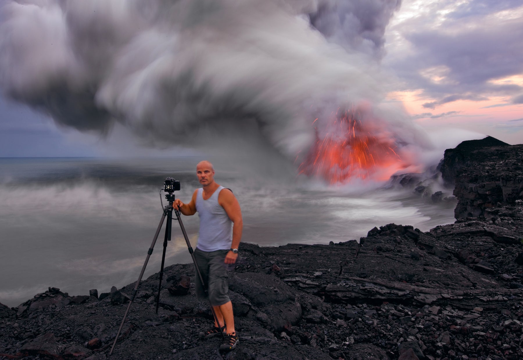 Portrait of Peter Lik with a tripod standing on black lava shores of Hawaii as hot lava pours into the Pacific creating plumes of thick gray smoke in the background