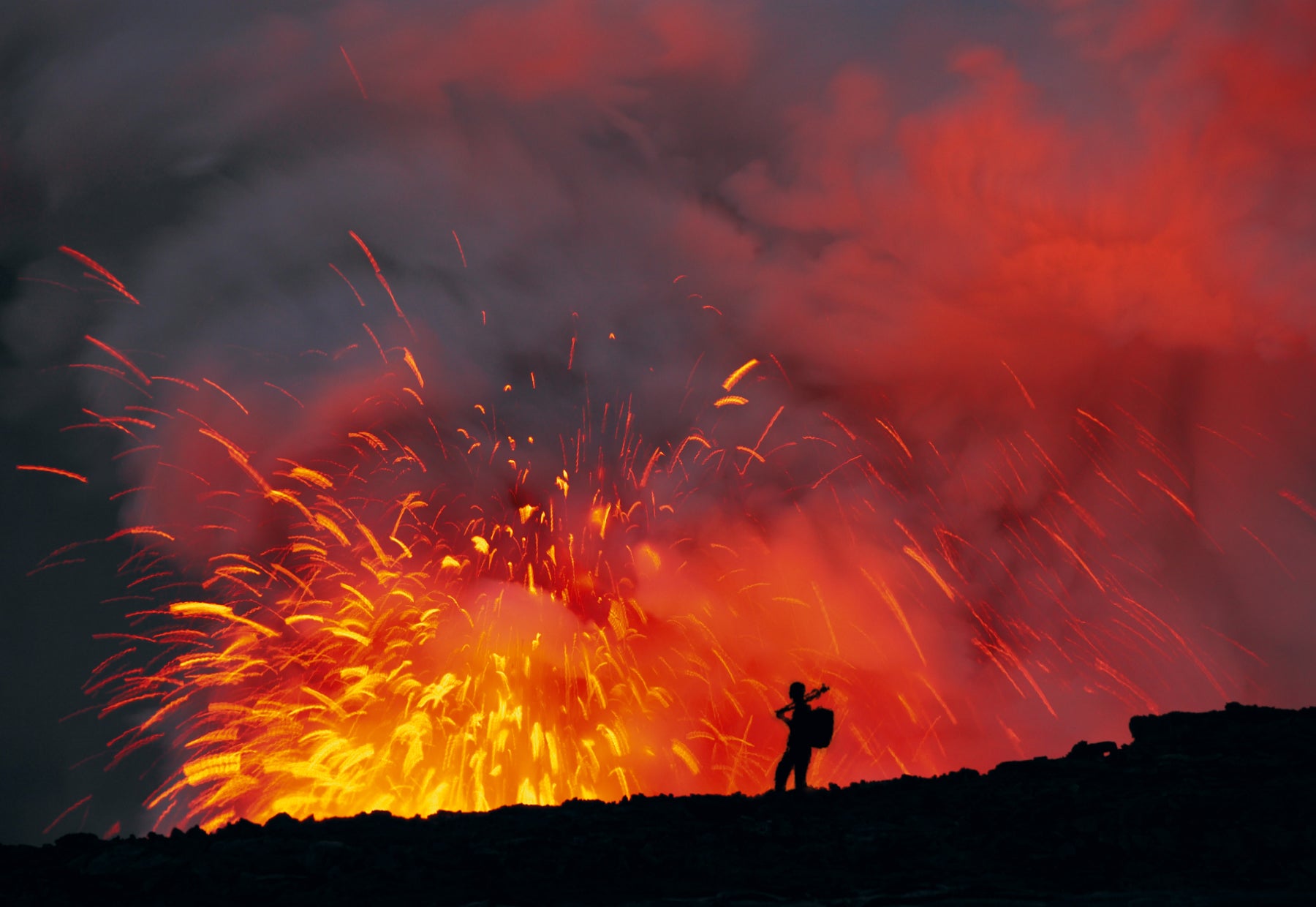 Silhouetted portrait of Peter Lik wearing a backpack and carrying a tripod traveling across black lava fields of Hawaii as explosions of lava light the smoke bright red