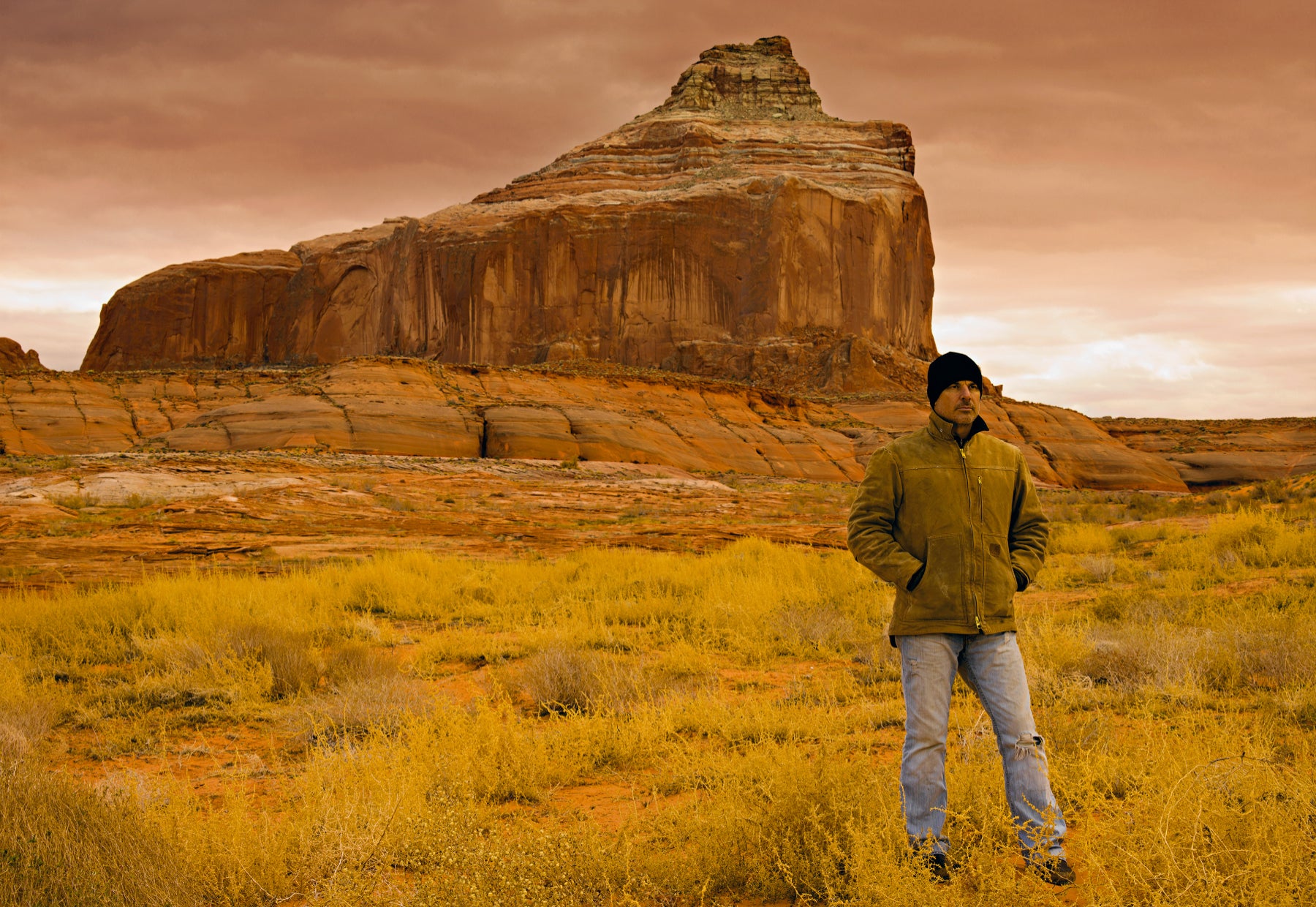 Portrait of Peter wearing a Carhart Jacket and beanie standing in the desert grass in front of towing weather carved stone cliffs