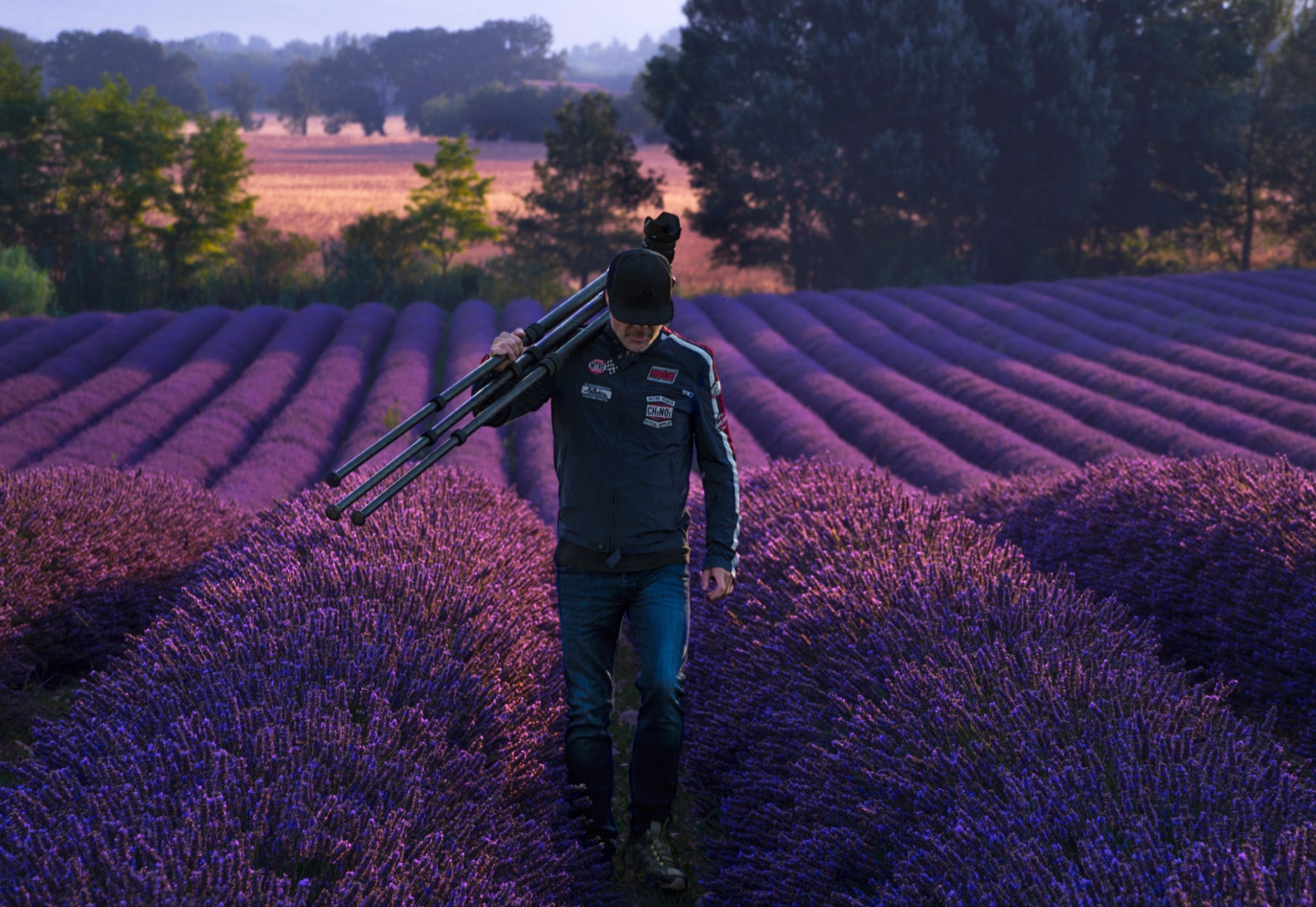 Portrait of Peter Lik in a baseball cap and leather motorcycle jacket walking with a tripod through the lavender fields of Valensole, France.