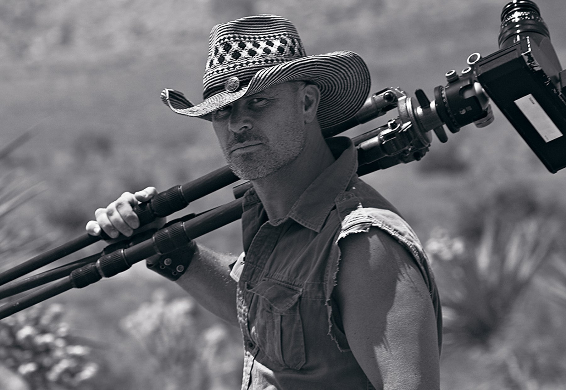 Stoic black and white portrait of Peter Lik wearing a cowboy hat and sleeveless shirt with a Linhoff 617 camera on a tripod resting on his shoulder in the desert of Red Rock Canyon, Nevada
