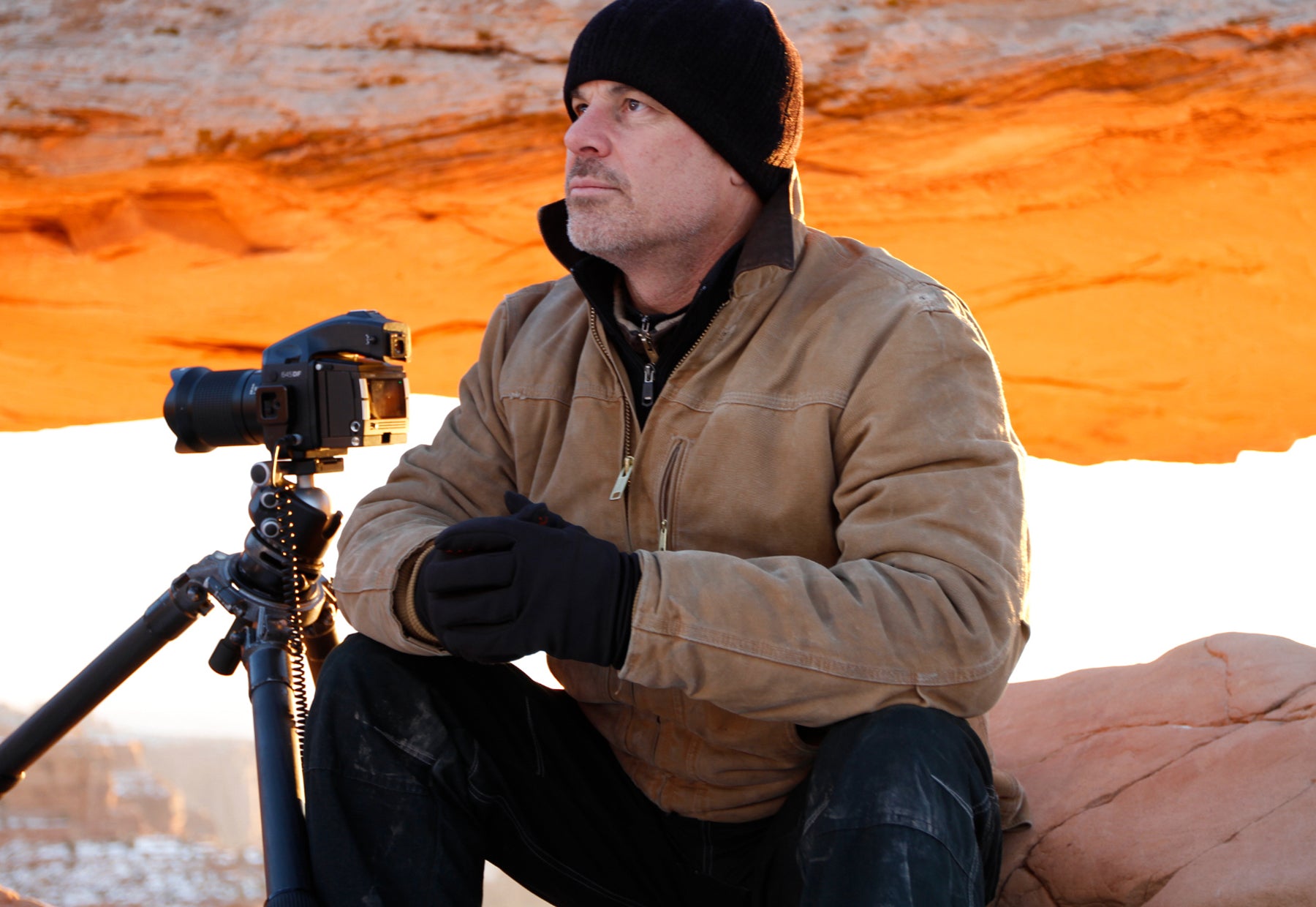 Portrait of Peter Lik wearing a Carhart jacket and beanie sitting with his camera and tripod in front of Mesa Arch at Canyonlands National Park in Utah during sunset