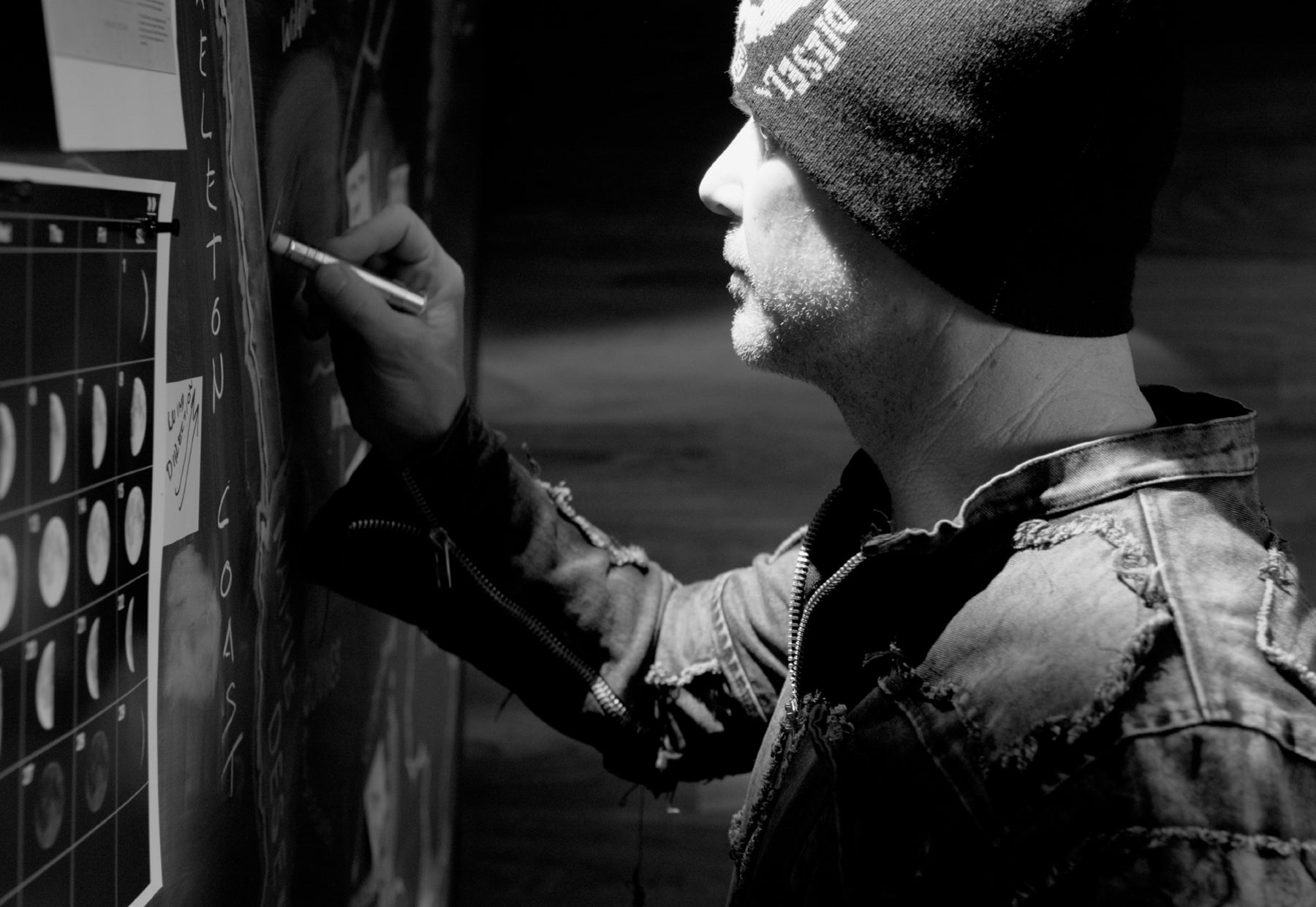Backlit black and white portrait of Peter Lik wearing a black leather jacket and beanie writing notes on a chalkboard as he researches new locations to photograph
