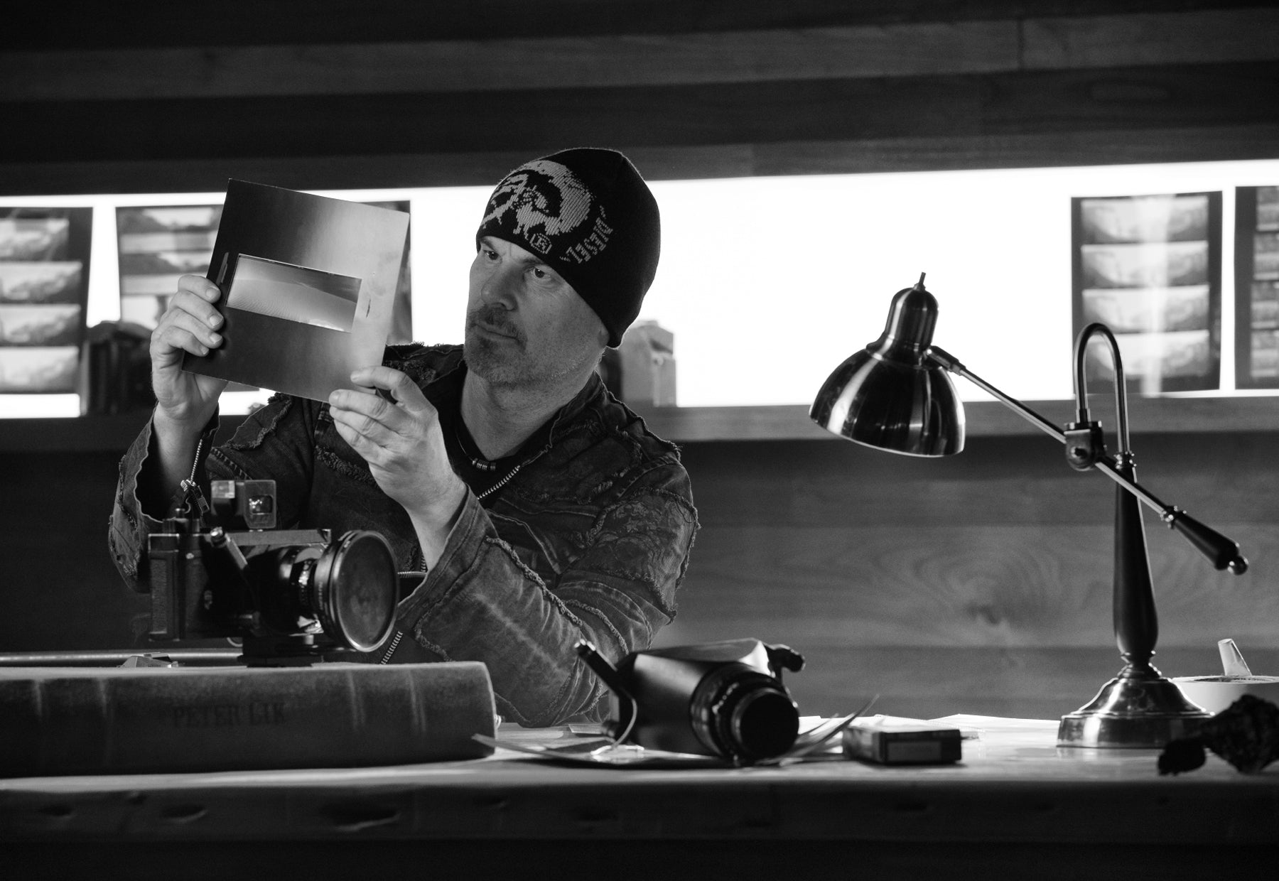 Backlit black and white portrait of Peter Lik wearing a black leather jacket and beanie sitting at a desk adorned with a Linhoff 617 III camera and copy of his 25th Anniversary Big Book