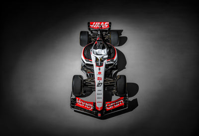 Photograph of the red, white, and black Haas F1 race car from above lit by a spotlight on the concrete floor of a airplane hanger.