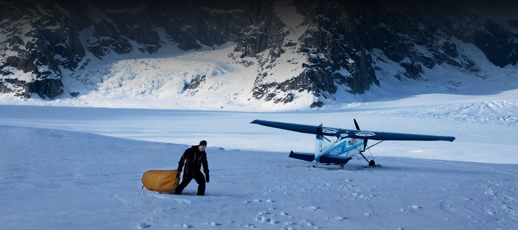 Portrait of Peter Lik walking in deep snow next to a blue and white bush plane in Alaska with the Denali Mountains in the background.