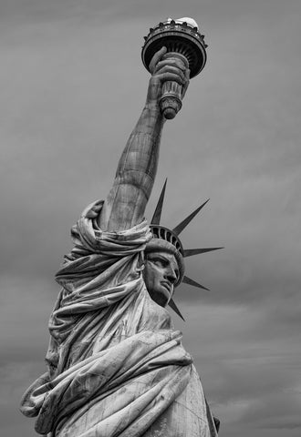 Black and white fine art photograph of the Statue of Liberty from below, highlighting her torch, crown, robe, and powerful expression beneath a dramatic sky.