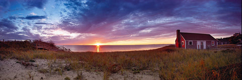 Sun setting on the ocean behind a red cottage on the grass covered beach of Cape Cod Massachusetts