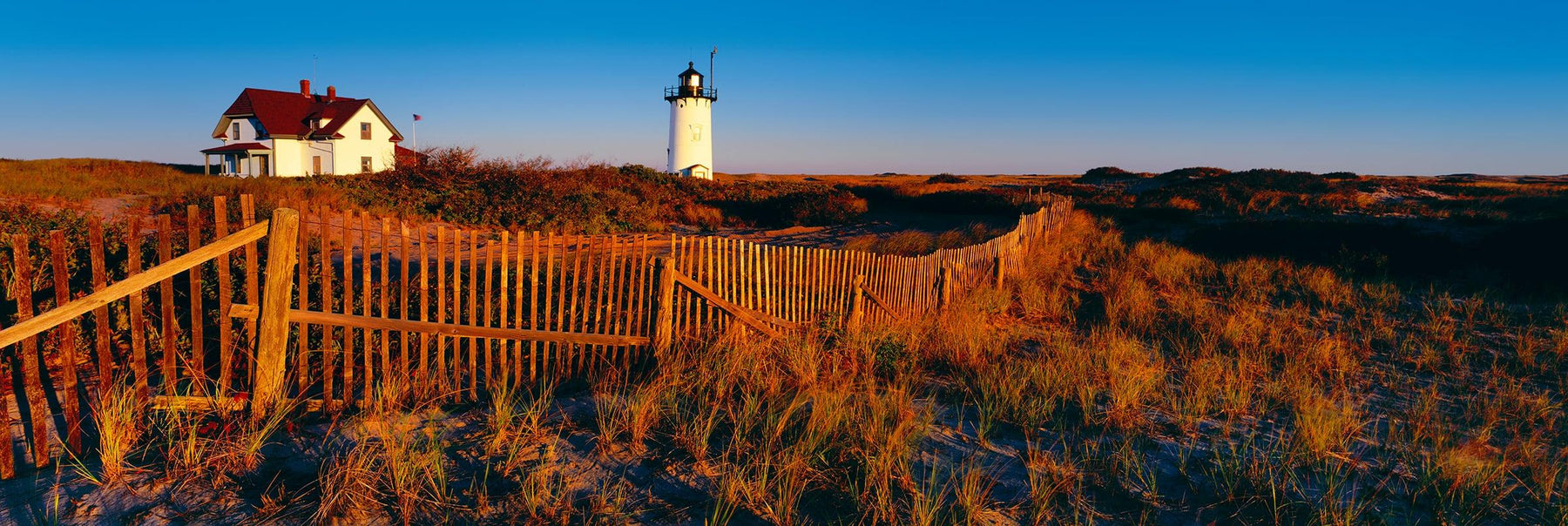 Wooden picket fence in front of a house and lighthouse on the brush covered coast of Cape Cod Massachusetts 