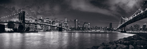 Black and white rocky rivers edge looking across to the lit up bridges leading into the New York cityscape  
