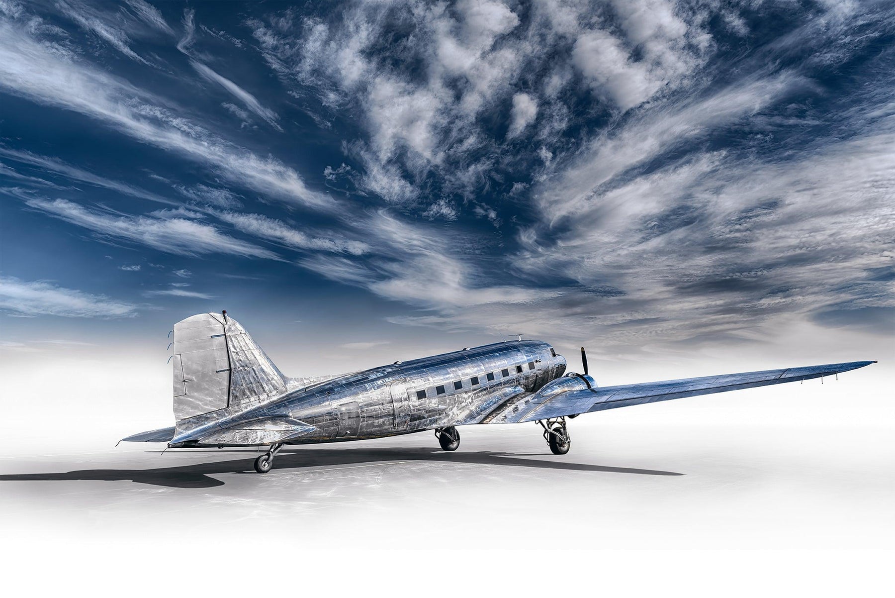 Silver DC-3 airplane with a white foreground and a cloudy blue sky above