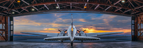 Silver DC-3 airplane in an open metal hanger with cloudy sky at sunset