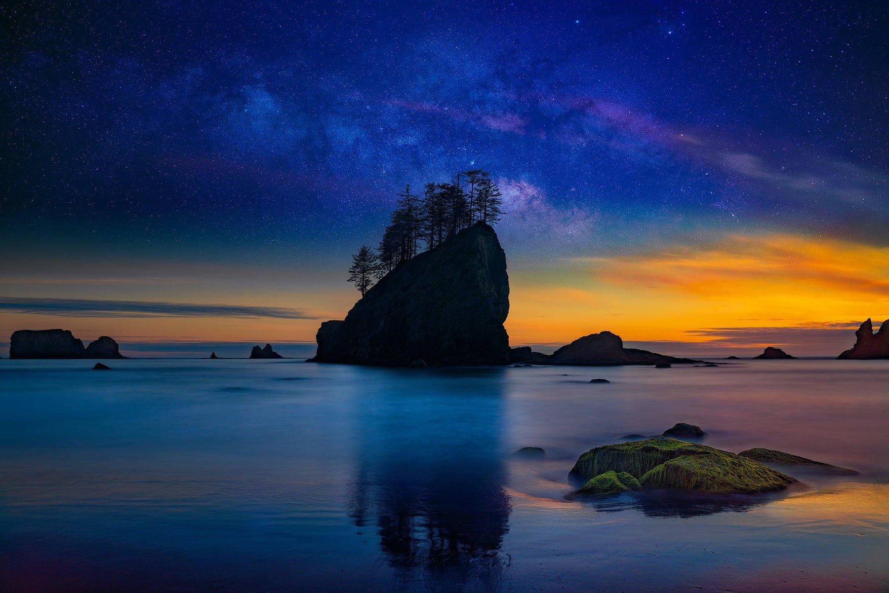 Silhouette of a sea stack covered with trees reflecting off the beach at sunset with the stars and milky way behind
