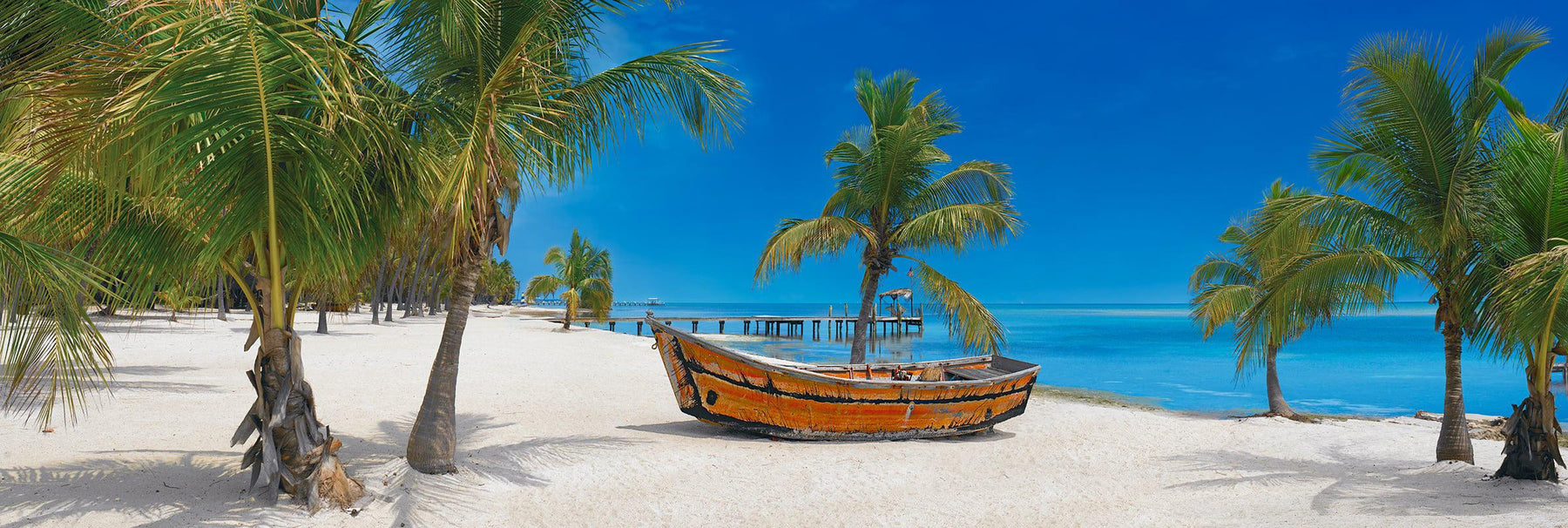 Old wooden boat sitting on a white sand beach full of palm trees with a jetty and the ocean in the background