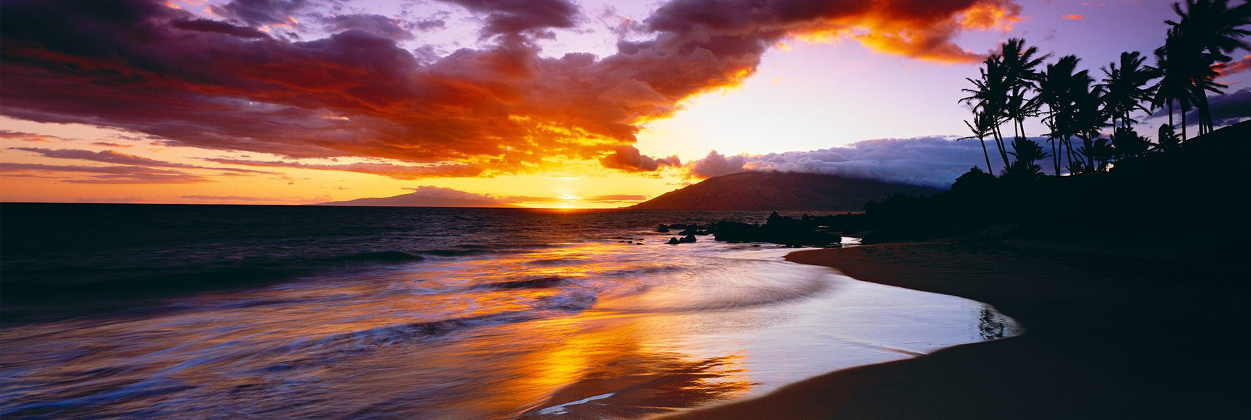 Waves crashing along the sand at Kihei Beach with the sun setting through the clouds in the background