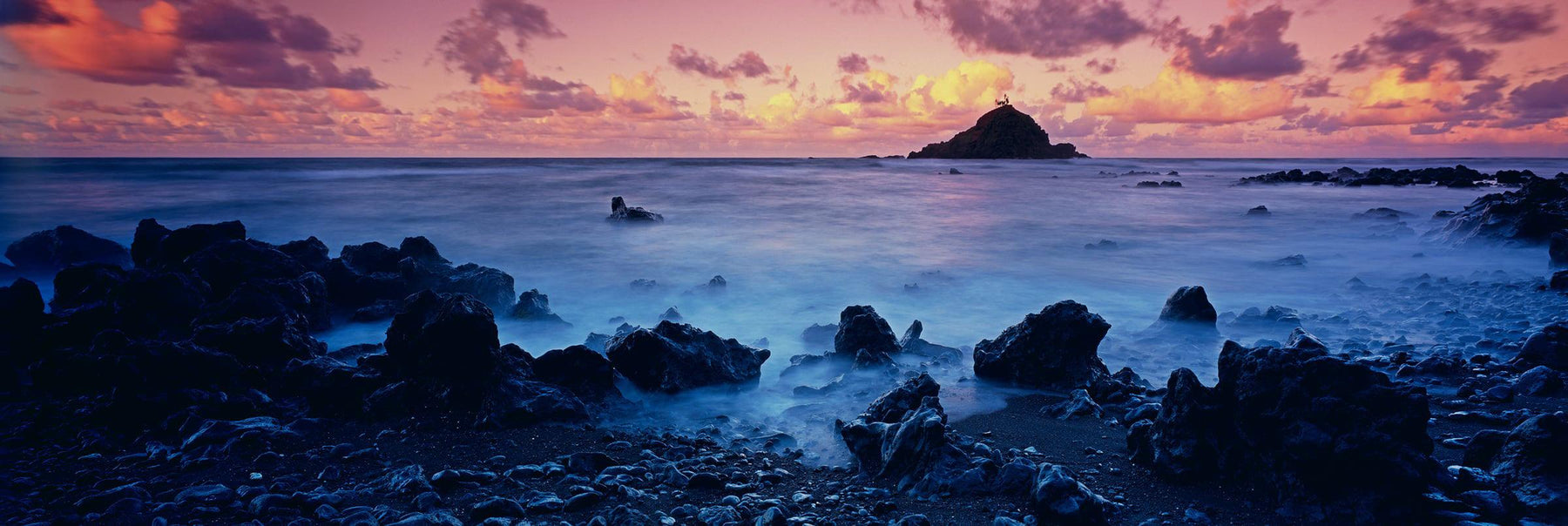 Waves washing onto a rocky Koki Beach with a distant island silhouetted in the background at sunrise