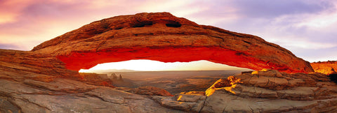 Stone arch glowing red from the sun rising over the stone peaks and and valley of Canyonlands National Park Utah