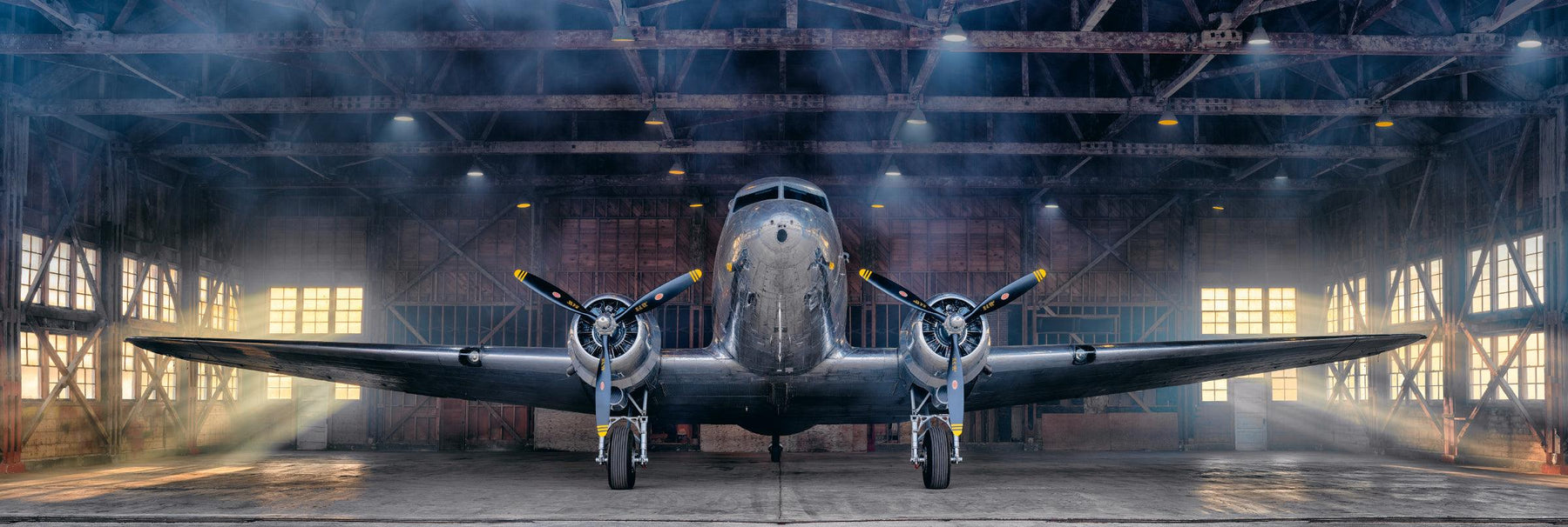 Silver DC-3 airplane in an open metal hanger with sun shining in the windows