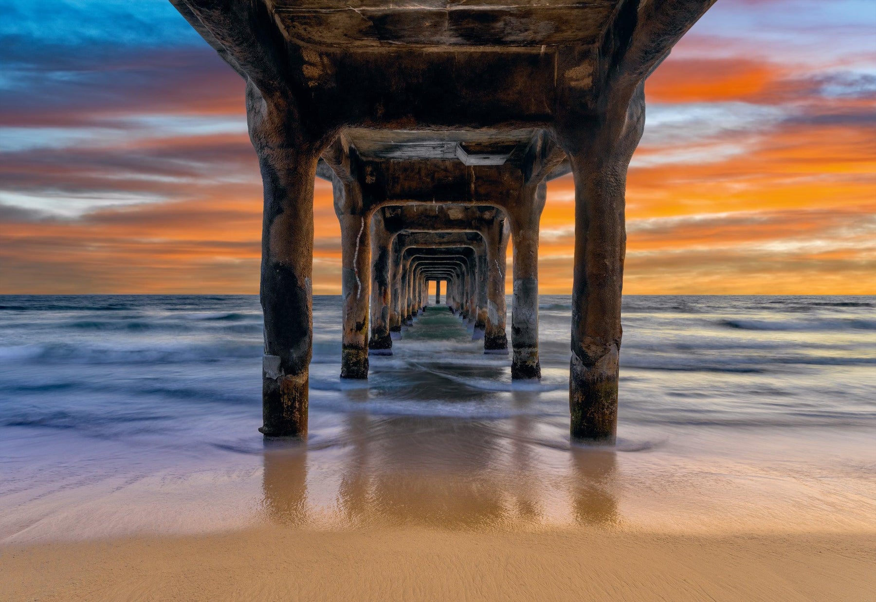 Waves crashing under a pier and onto the sand during a cloudy sunset